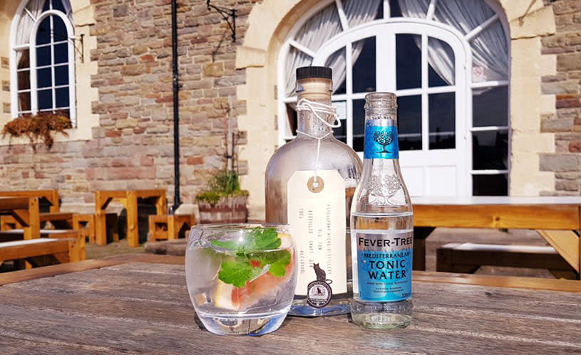 A bottle of gin, a bottle of tonic and a gin and tonic in a glass on a wooden table outside The Pump House - Credit Visit Bristol