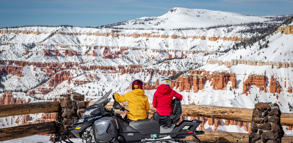 Two people dressed in bright yellow and pink sit on a parked snowmobile behind a wooden fence staring out at sweeping red rock formations dusted in snow.