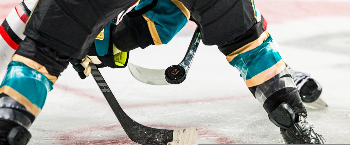 Close-up of two hockey players in action during a face-off. They wear teal and black gear. The puck is suspended between their crossed sticks on ice.