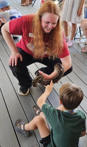 A trained library volunteer holds a snake on her arm and extends it to a student so that he may pet it safely.