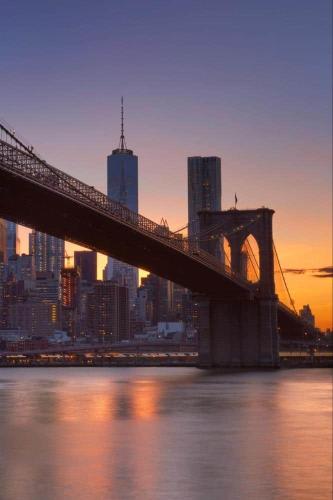 Brooklyn Bridge spanning the East River at dusk, with One World Trade Center and the illuminated Manhattan skyline glowing against a warm orange and purple sunset sky