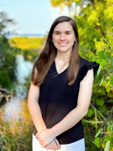 Portrait of Kristen smiling outdoors, standing in front of lush greenery with water and blue sky in the background. She is wearing a black blouse and white pants.