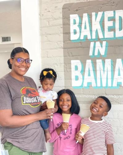 A mother and her three children stand in front of a painted message on a brick wall that reads "BAKED IN BAMA." Two of the children hold ice cream cones, and the mother holds the ice cream come for her youngest, who she holds in her other hand.