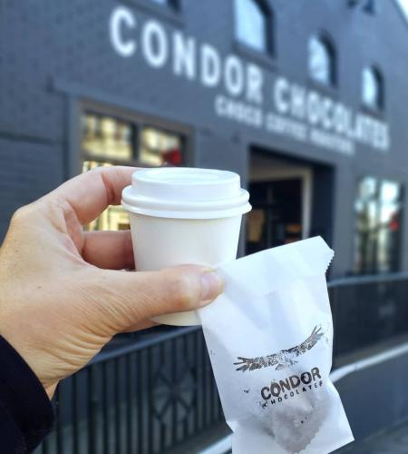 A hand holds a to-go cup and small Condor Chocolates bag in front of Condor's new downtown store front.
