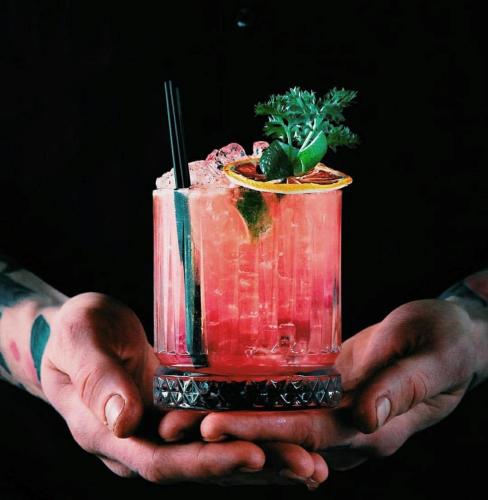 The Night Owl pink cocktail with garnish and straw being held in two hands with black background