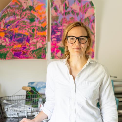 Woman smiles and stands in front of completed artwork hanging on the wall. She is wearing a white shirt and black-framed glasses.