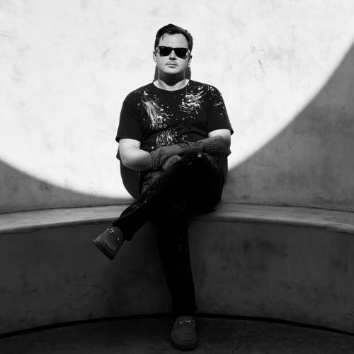 black and white photo. Man wears black sunglasses, dressed in black, and crosses his arms.He leans on a concrete wall, centered within a circular shadow.