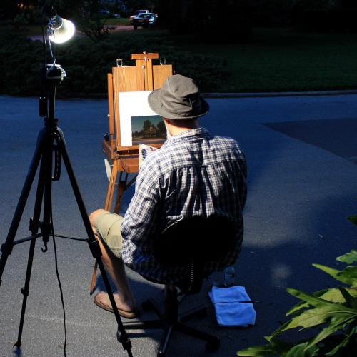 man sits and paints a photo leaning on an easel in a dark space. THere is a single light shining on his work.