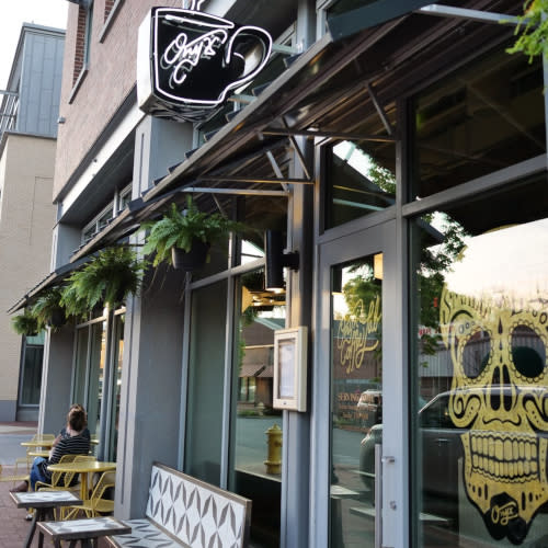 An image of the entrance of Onyx Coffee Lab at 100 NW 2nd Street in Bentonville, AR. Featuring a decorative neon sign that spells Onyx, a gold sugar skull illustration on the window, and an outdoor seating area with hanging baskets of green ferns.