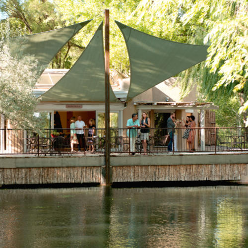 A group of people socialize on a shaded patio overlooking a tranquil pond, surrounded by lush greenery.