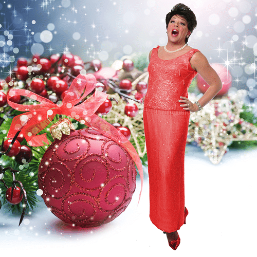 Judy Show at the Purple Room: A person in a festive red outfit expressing excitement beside a large Christmas ornament with holiday decorations in the background.