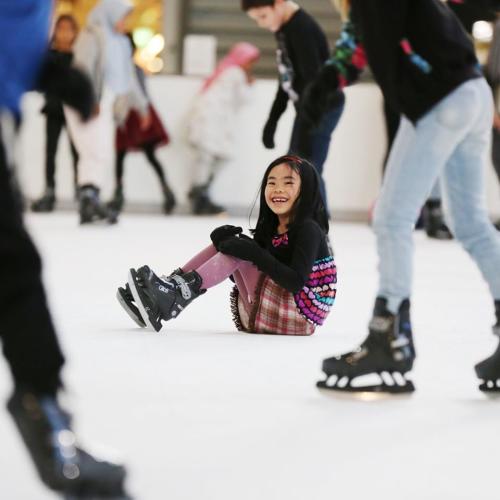 A little girl sitting in the middle of a skating rink while people skate around her