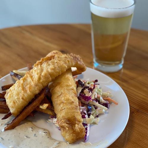 Fried Fish over Slaw on a white plate sitting on a wooden table with a glass of pale frothy beer