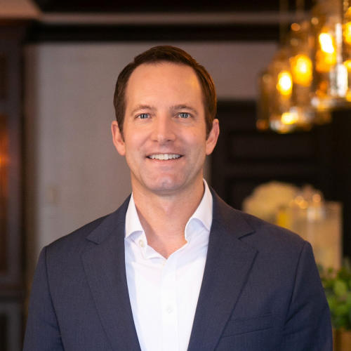A headshot of VTW Director Hank Reilly. He's 30s-40s with short, dark brown hair and blue eyes. He's wearing a white button-down shirt and navy summer blazer, standing in a restaurant with upscale golden lights.