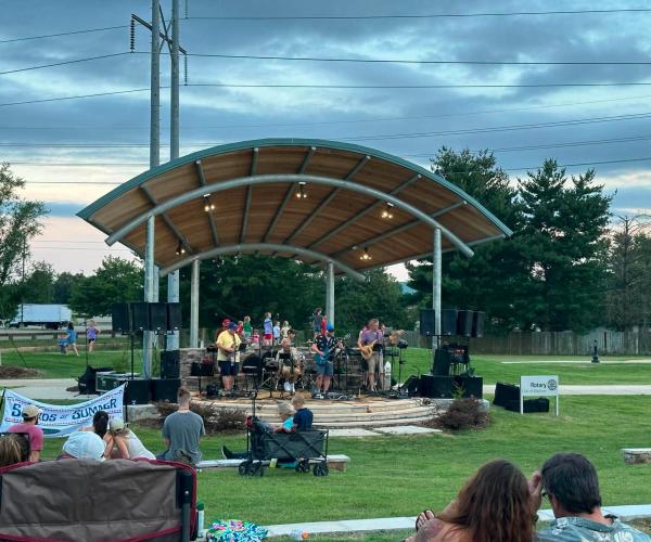 A live band performs under the pavilion during the Sounds of Summer concert series in Madison. Families and friends relax on the lawn in chairs and blankets while enjoying an evening of live music in the park beneath a cloudy summer sky.