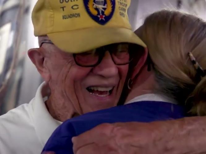 Older military veteran wearing a yellow cap smiles while embracing a woman during an emotional reunion moment, captured in a close-up setting.