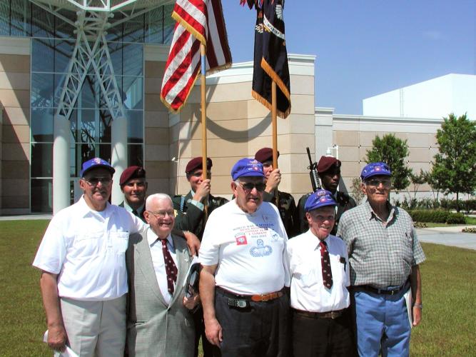 A group of veterans wearing matching caps poses for a photo outside a modern building, with color guard members holding U.S. and military flags behind them.