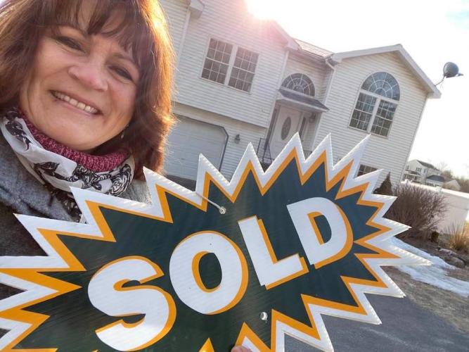 A woman smiling and holding a big green SOLD sign in front of a cream-colored house.