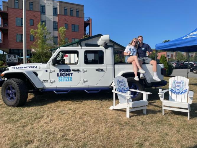 White Jeep with Bud Light logo, two people posing off the side of the bed of truck, two Adk chairs in front