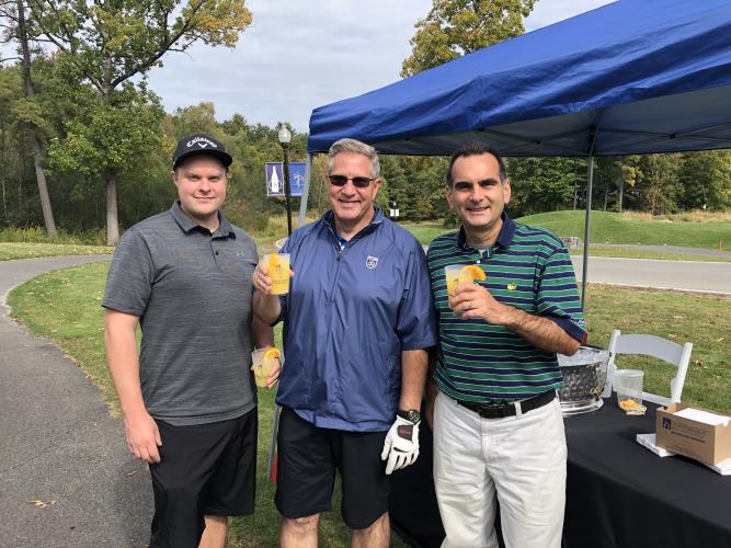 Three golfers posing with their drinks in front of a blue tent.