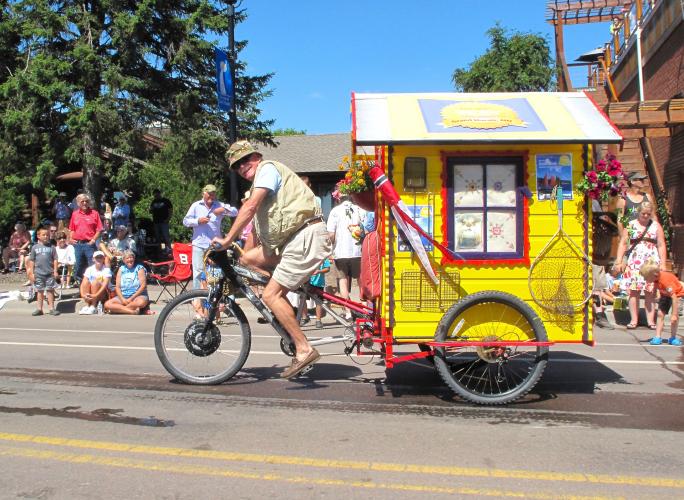 An individual riding a bicycle pulling a small house down the street in a parade