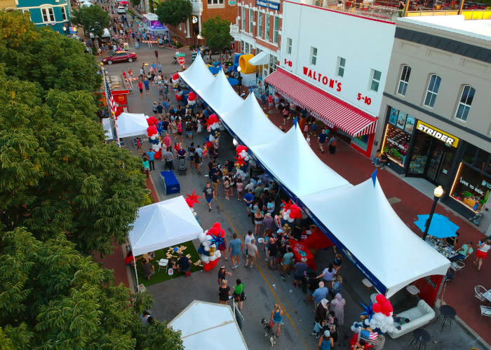 An aerial photo of the Bentonville square with green trees in the foreground, pedestrians strolling the street alongside tents, and the Walton 5 & 10 in the background.