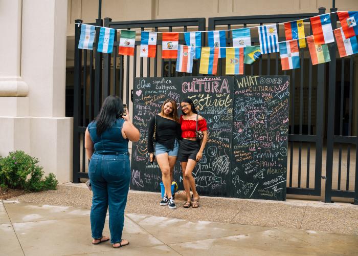Two attendees pose for a photo in front of a colorful community message wall and international flags at the CulturA Festival at The Orion Amphitheater in Huntsville.