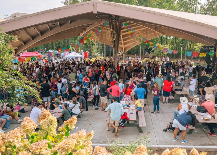 Large crowd gathers beneath the pavilion during the CulturA Festival at The Orion Amphitheater in Huntsville, surrounded by colorful decorations and vendor booths.