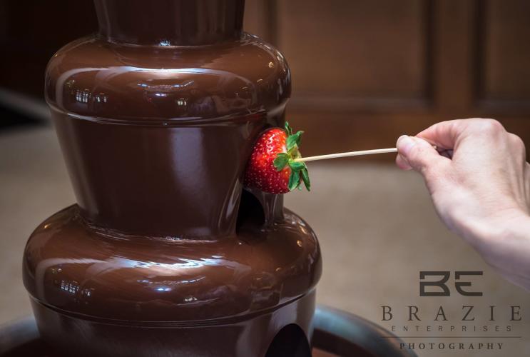 Strawberry on a stick being dipped into a multi-tiered chocolate fountain.