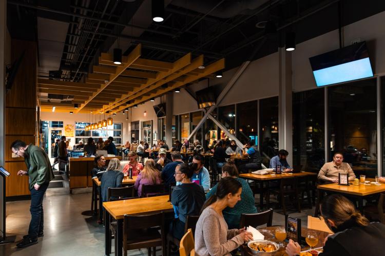 A bustling restaurant interior with wooden tables and a modern design. Diners enjoy meals and drinks under warm lighting, creating a lively atmosphere.