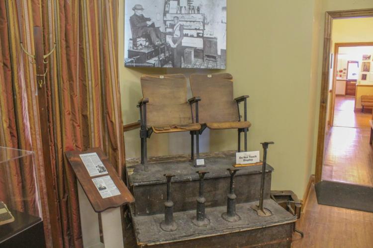 Rabbit Dickerson's original shoe shining station - wooden seats with metal supports, displayed in a museum setting. A black-and-white photo of a man in a shop hangs above.