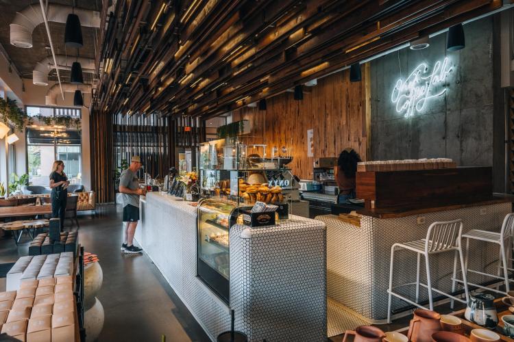 A modern café interior featuring a sleek counter with pastries, a barista serving customers, and stylish seating. Warm lighting and wooden accents create a welcoming atmosphere.