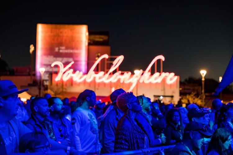Crowd at a nighttime event, illuminated by vibrant blue lights, with a prominent "You belong here" neon sign in the background.