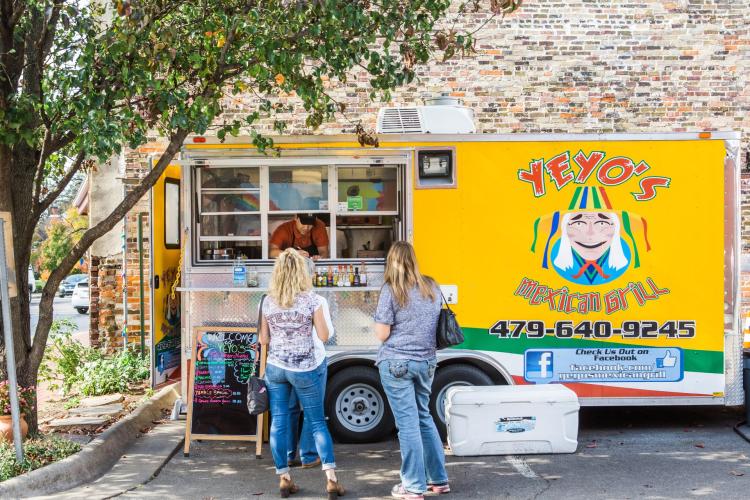 A vibrant yellow food truck with "Yeyo's Mexican Grill" signage, serving customers in a lively outdoor setting. Two women stand in line, engaging with the vendor.