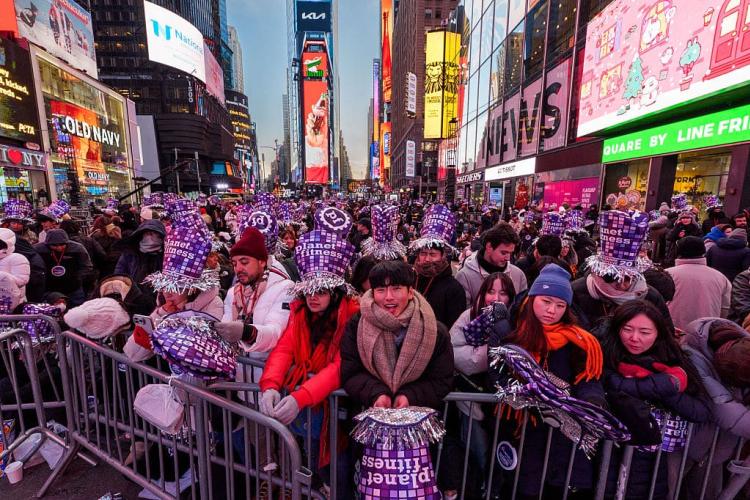 A massive crowd packed behind barricades in Times Square on New Year's Eve, wearing purple Planet Fitness party hats beneath the glow of illuminated billboards.