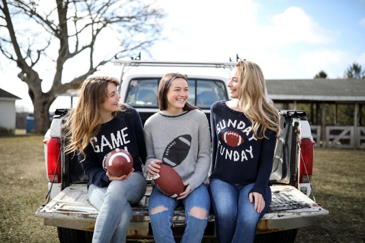 Three young women sitting on the tailgate of a truck holding footballs and wearing football themed sweaters