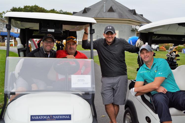Foursome posing with two men in a golf cart, one standing in between carts and one hanging out the side of the other