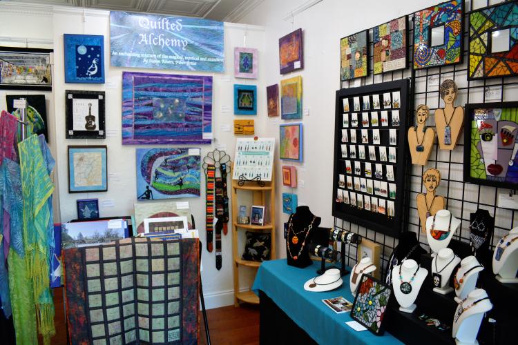 Corner interior shot with colorful quilts and jewelry hanging from the walls and displayed on tables.