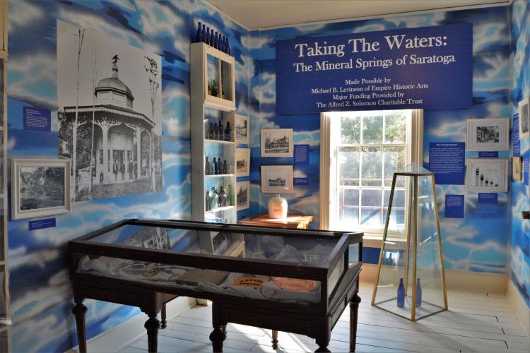 Taking the Waters exhibit, overview of the room with blue multi-colored walls and a glass covered display table