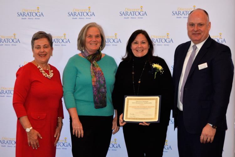 Group of four people standing in front of Discover Saratoga's step and repeat while accepting hometown ambassador award at Saratoga Springs City Center