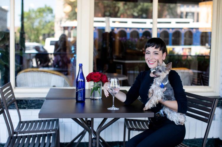 A woman drinking a glass of Saratoga water while holding her small dog on the The Blue Hen Patio in downtown Saratoga Springs NY