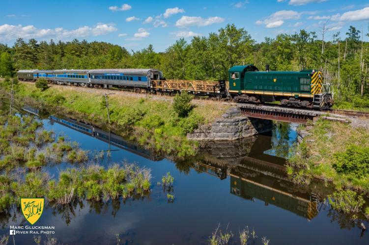 Full length of train riding alongside water surrounded by wooded area and fields