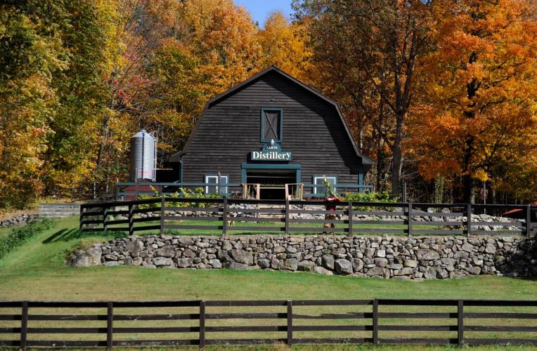 Outdoor shot of brown barn at Springbrook Hollow Farm with two rows of brown fencing and a stone wall in front and autumn foliage behind it.