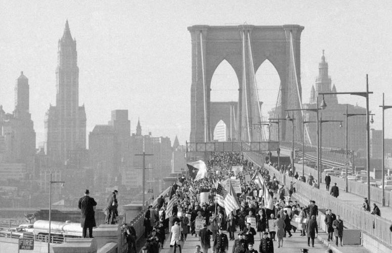 Black-and-white photograph of a large crowd of demonstrators marching across the Brooklyn Bridge's pedestrian walkway toward Manhattan, carrying American flags and protest signs, with the Lower Manhattan skyline — including the Woolworth Building — visible in the background.