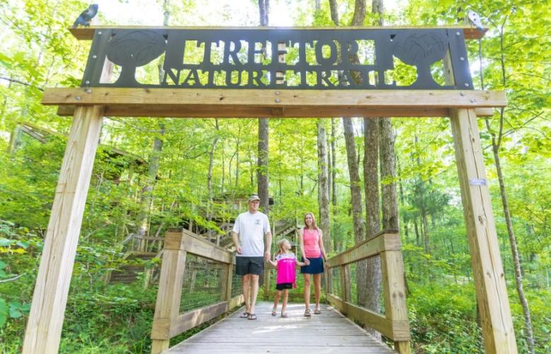 A father, mother, and daughter walk across a wooden bridge in the middle of a lush, green forest. The sign hanging over the bridge reads "TREETOP NATURE TRAIL."