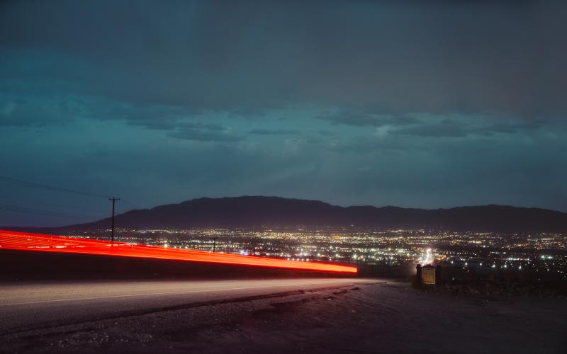 A long exposure of a car driving down Nine Mile Hill at night, leaving a red streak of light. The city lights of Albuquerque are illuminated below the shadowy Sandia mountains.