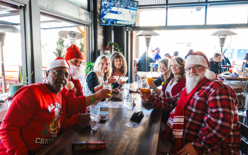 A group wearing festive holiday attire and santa hats sit at a bar-top table holding their drinks up in the air smiling.