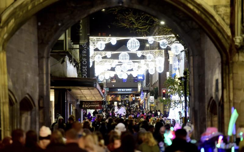 A crowd of people moving along a high street at night with large Christmas lights decorations glowing brightly overhead.