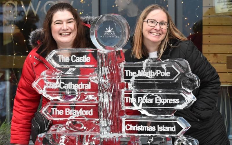 Two women stood posing with an ice sculpture of a signpost.