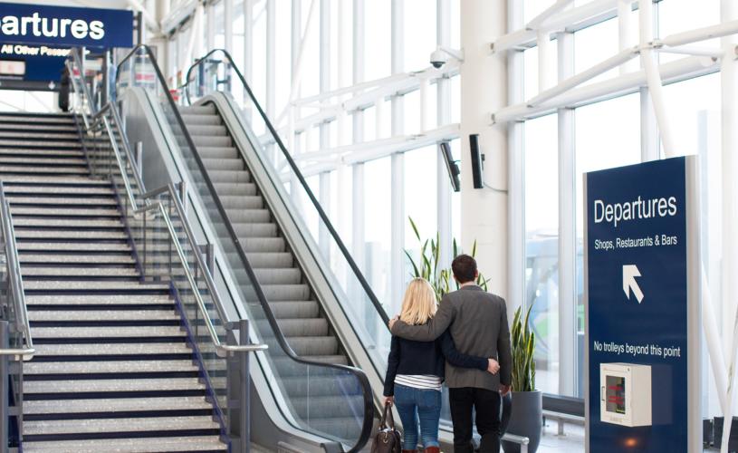 2 people stepping on a escelator going up to the departure lounge at Bristol Airport - Credit Bristol Airport
